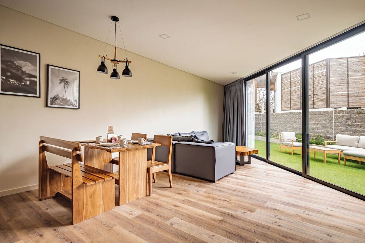 A modern living room with a wooden dining table, grey sofa, and large sliding glass doors leading to a garden.