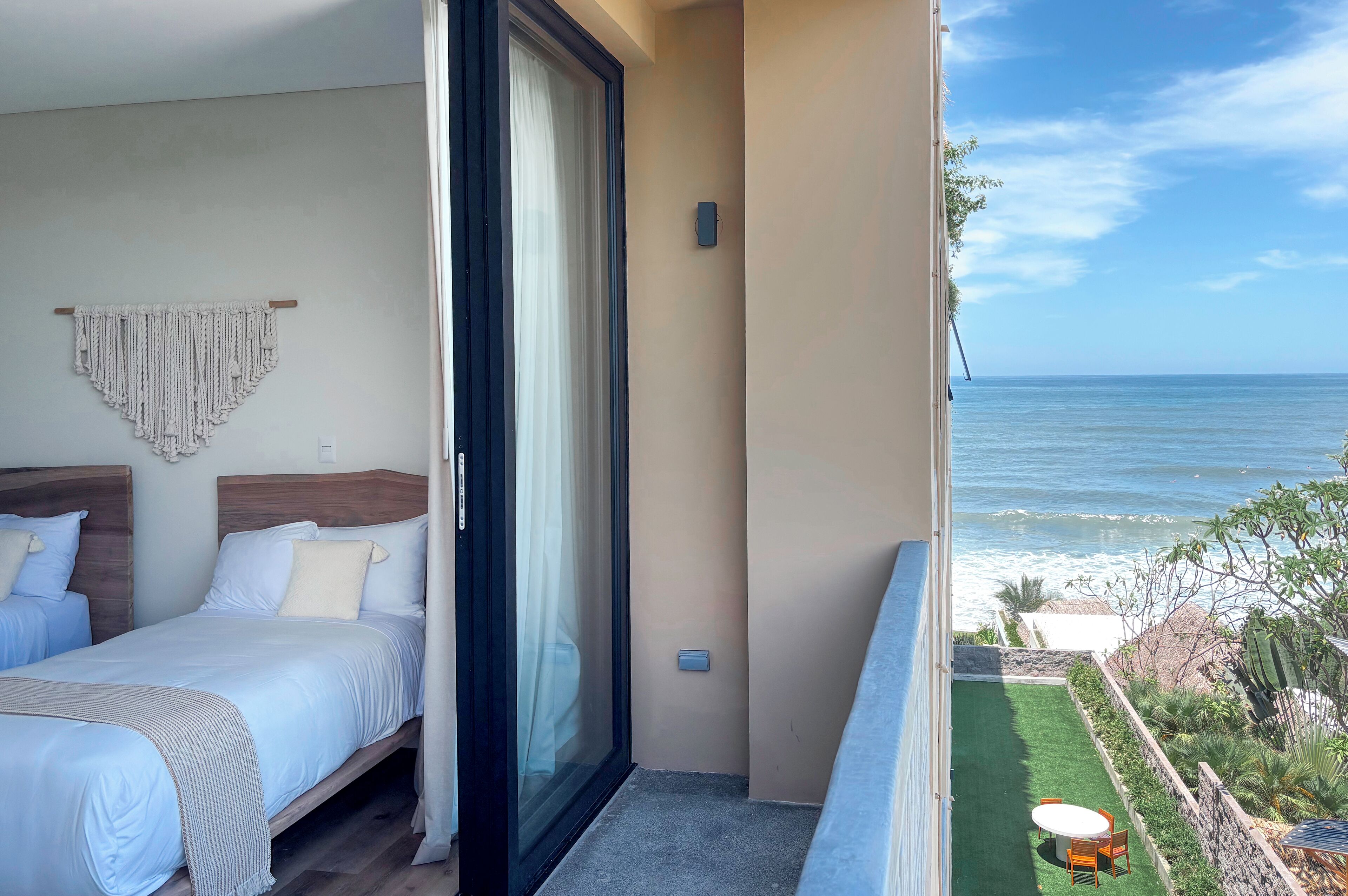 Ein Balkon mit Blick auf den Ozean, ein Bett mit weißer Bettwäsche und ein Wandbehang aus Makramee.