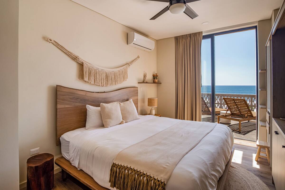 A bedroom with a large bed, a ceiling fan, an air conditioning unit, a bedside lamp, a wooden nightstand, a wooden stool, a macrame wall hanging, a shelf with decorative items, and a balcony with a view of the ocean.