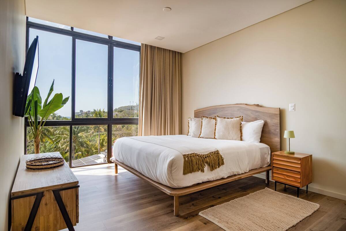 A bedroom with a large window, a bed with white bedding, a wooden headboard, a bedside table with a lamp, and a woven basket on a wooden nightstand.