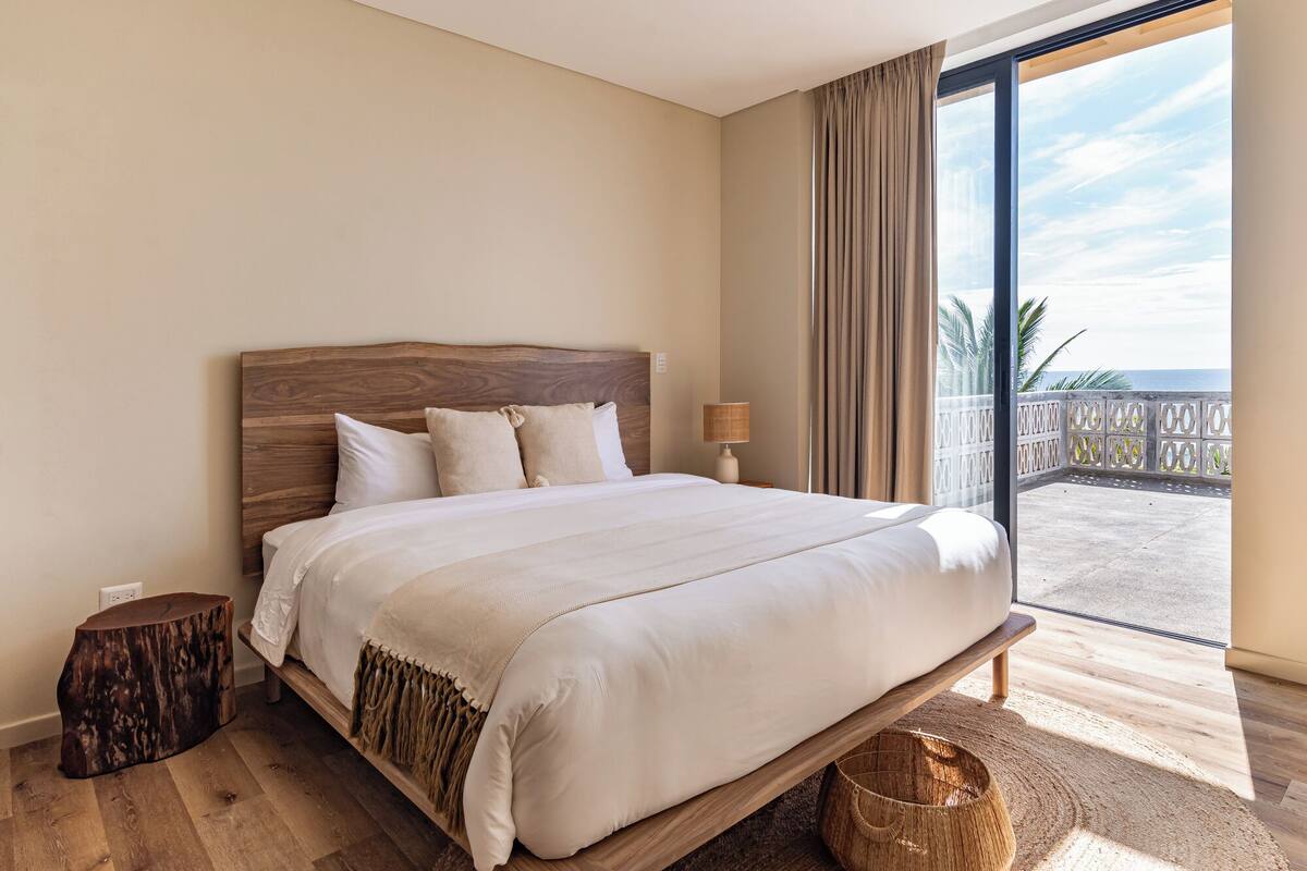A bedroom with a large bed, a nightstand, a lamp, and a view of the beach through a large window.