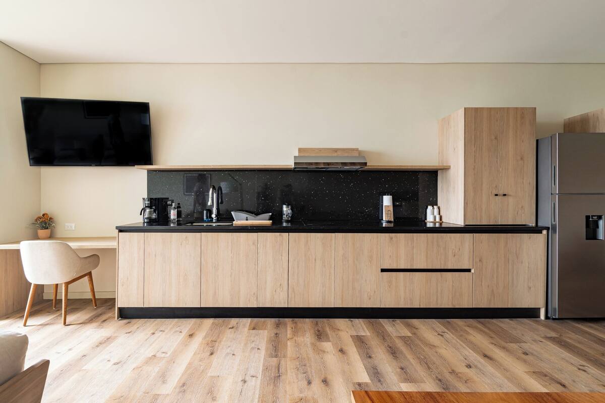 A modern kitchen with a black countertop, wooden cabinets, a stainless steel refrigerator, and a flat-screen TV mounted above the counter.