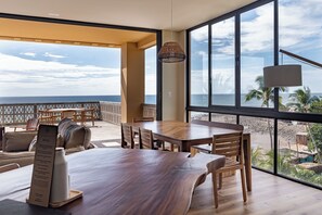 Un vaste espace dédié à la salle à manger, avec une grande table et des chaises en bois, donnant sur une plage bordée de palmiers et sur l’océan.