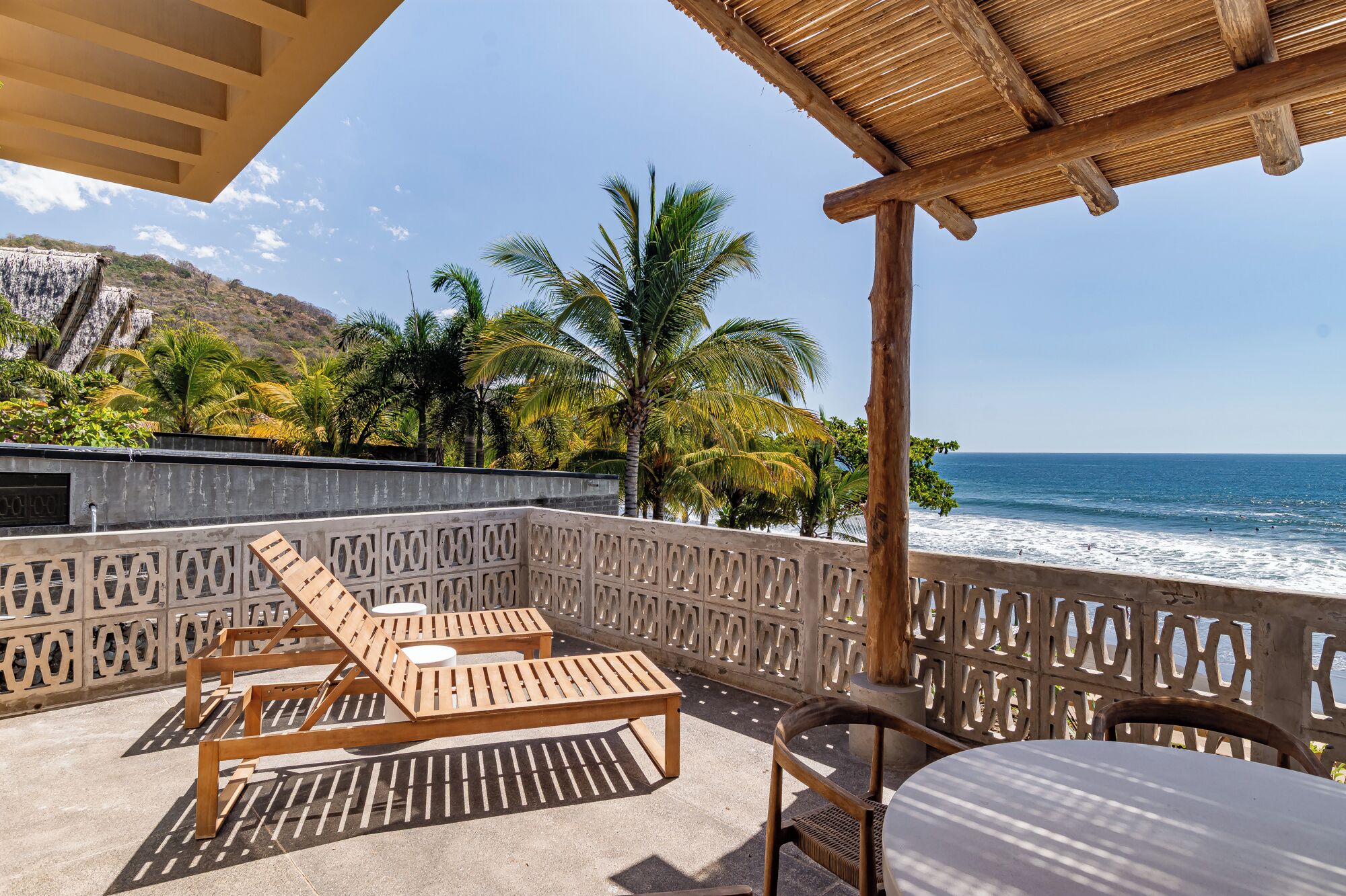 Un balcone con poltrone da relax in legno e un tavolo, con vista su una spiaggia con palme e l'oceano.