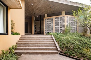 Une série de marches en béton menant à l’entrée d’un bâtiment, entourées de verdure et d’un mur décoratif orné de motifs géométriques.