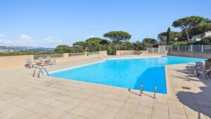 A rectangular swimming pool with clear blue water, surrounded by a tiled deck. There are lounge chairs and a small staircase leading into the pool. The area is enclosed by a low white fence, and trees and buildings are visible in the background. - Le Mazet aux Cactus - Maison avec piscine partagée (Cogolin)