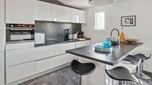 A modern kitchen with white cabinets, a black countertop, and a central island with bar stools. - Le Mazet aux Cactus - Maison avec piscine partagée (Cogolin)
