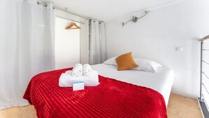 Et hotelværelse med en seng, hvide gardiner, et rødt tæppe, to håndklæder, en brun pude og en trægulv.