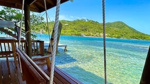 A wooden swing chair hanging from ropes on a wooden deck overlooking clear blue water and a green hillside. - Fork Tail Lodge  Secluded 3BR Waterfront Retreat (José Santos Guardiola)