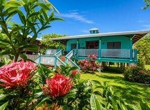 A turquoise house with a porch, surrounded by lush greenery and flowering plants.