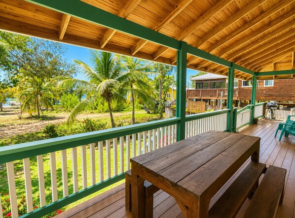 A wooden deck with a bench, a white picket fence, and a view of a beach and palm trees.