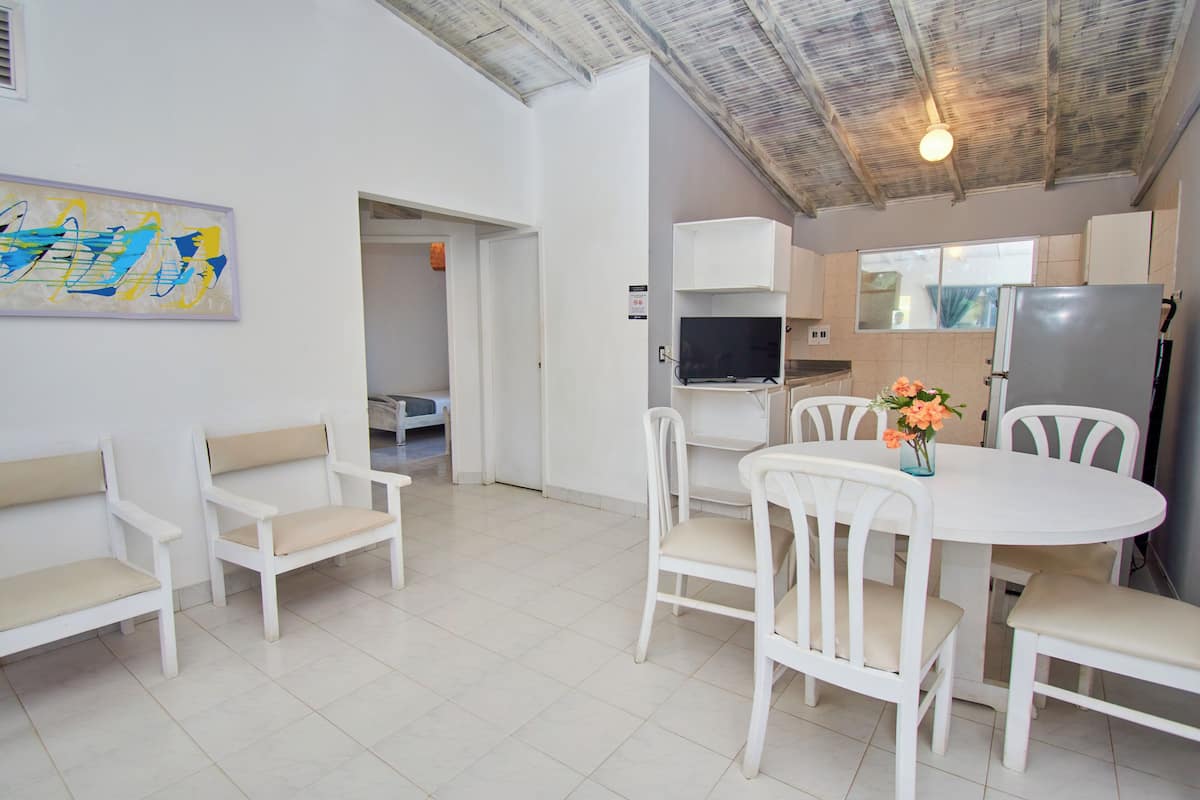 Une cuisine moderne avec des armoires blanches, un four encastré et un réfrigérateur. On y trouve un coin repas avec une table et des chaises blanches, ainsi qu’un tableau accroché au mur.