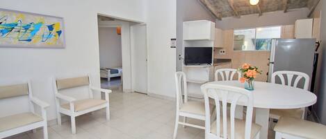 A modern kitchen with white cabinets, a built-in oven, and a refrigerator. There is a dining area with a white table and chairs, and a painting on the wall.