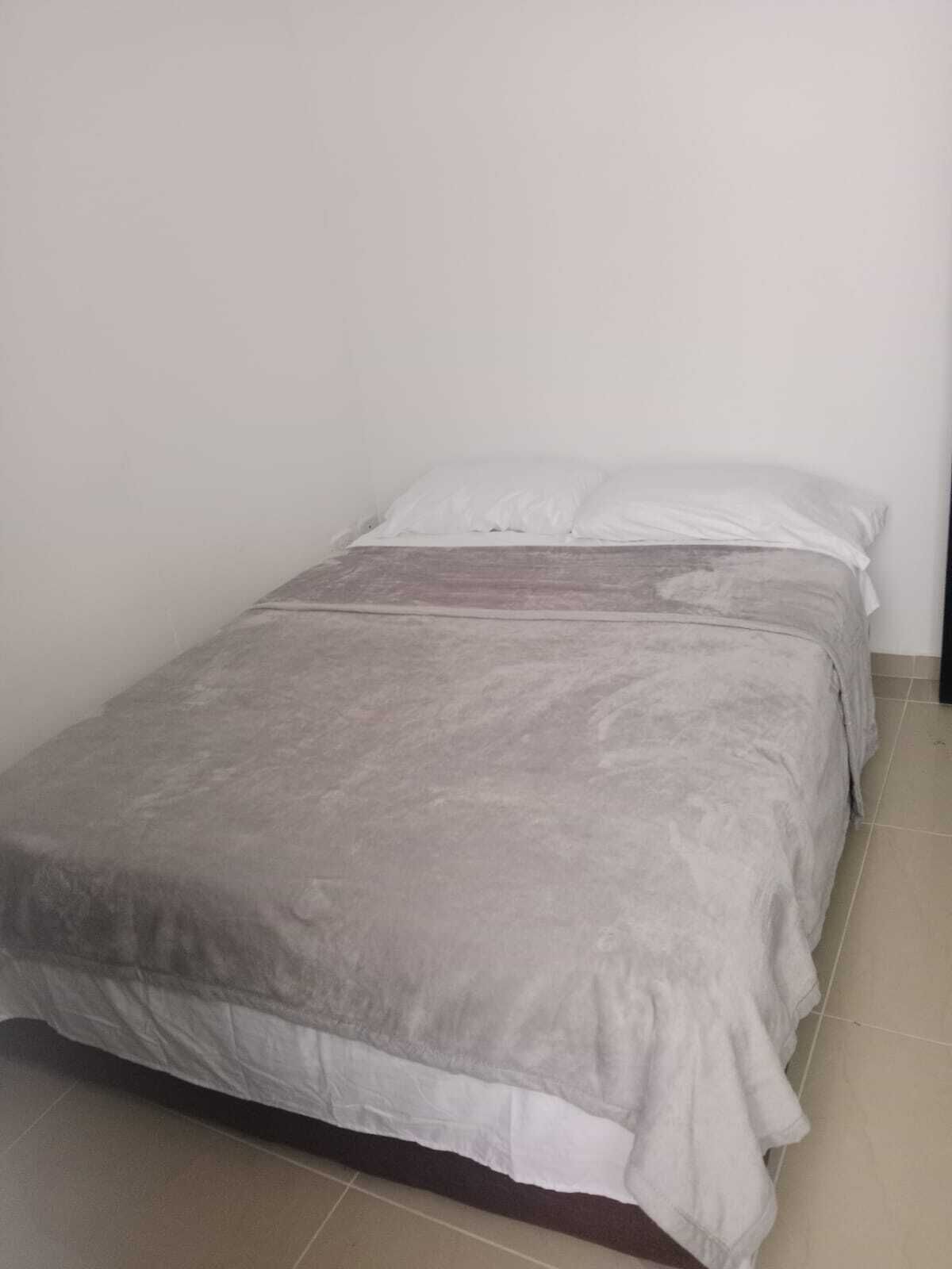 A small, single-bed room with a grey bedspread and white pillows. The room has plain white walls and a tiled floor.