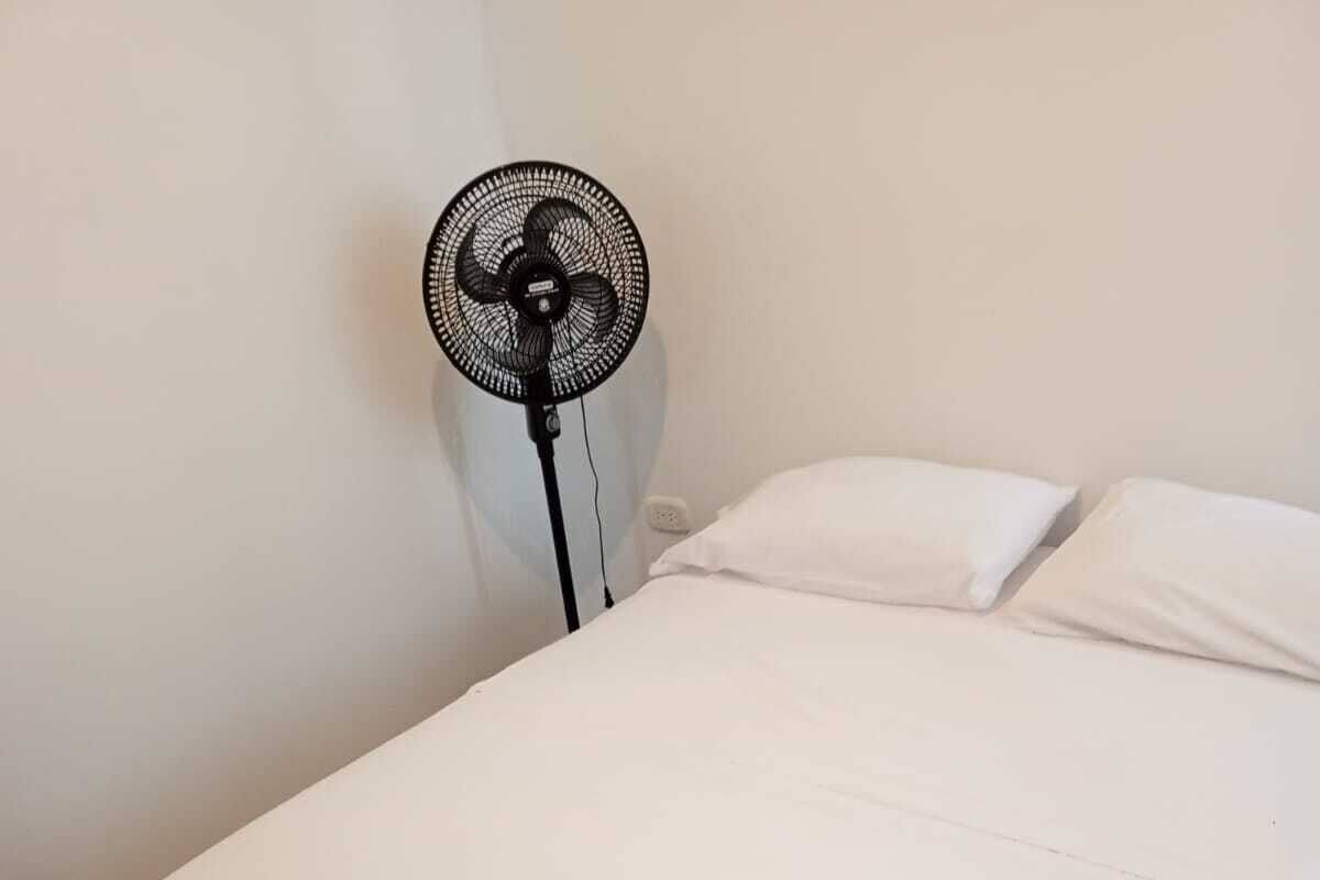 A single bed with white bedding in a room with a white wall and a standing fan.