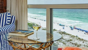 Una habitación con vistas a la playa, una mesa de vidrio con un libro y una copa de vino, y muebles a rayas azules y blancas.