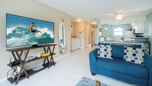 A living room with a TV, a blue sofa, and a kitchen in the background. - Miramar Beach | Walk to Beach & Shops (Miramar Beach)