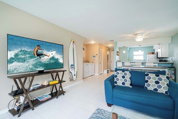 A living room with a TV, a blue sofa, and a kitchen in the background.