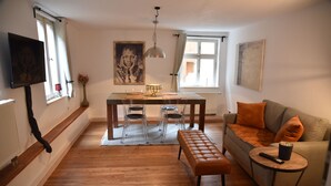 A living room with a wooden floor, a sofa with orange cushions, a round coffee table, a TV mounted on the wall, and a dining table with chairs. - The small guest house in the heart of the historic old town of Dinkelsbühl. (Dinkelsbühl)