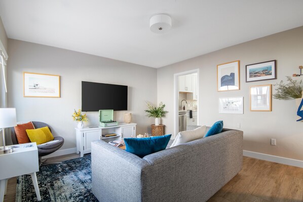 A modern living room with a grey sofa, a small white desk, a flat-screen TV, and a kitchen area visible through an open door. - Seashore Suites by AvantStay (La Jolla)