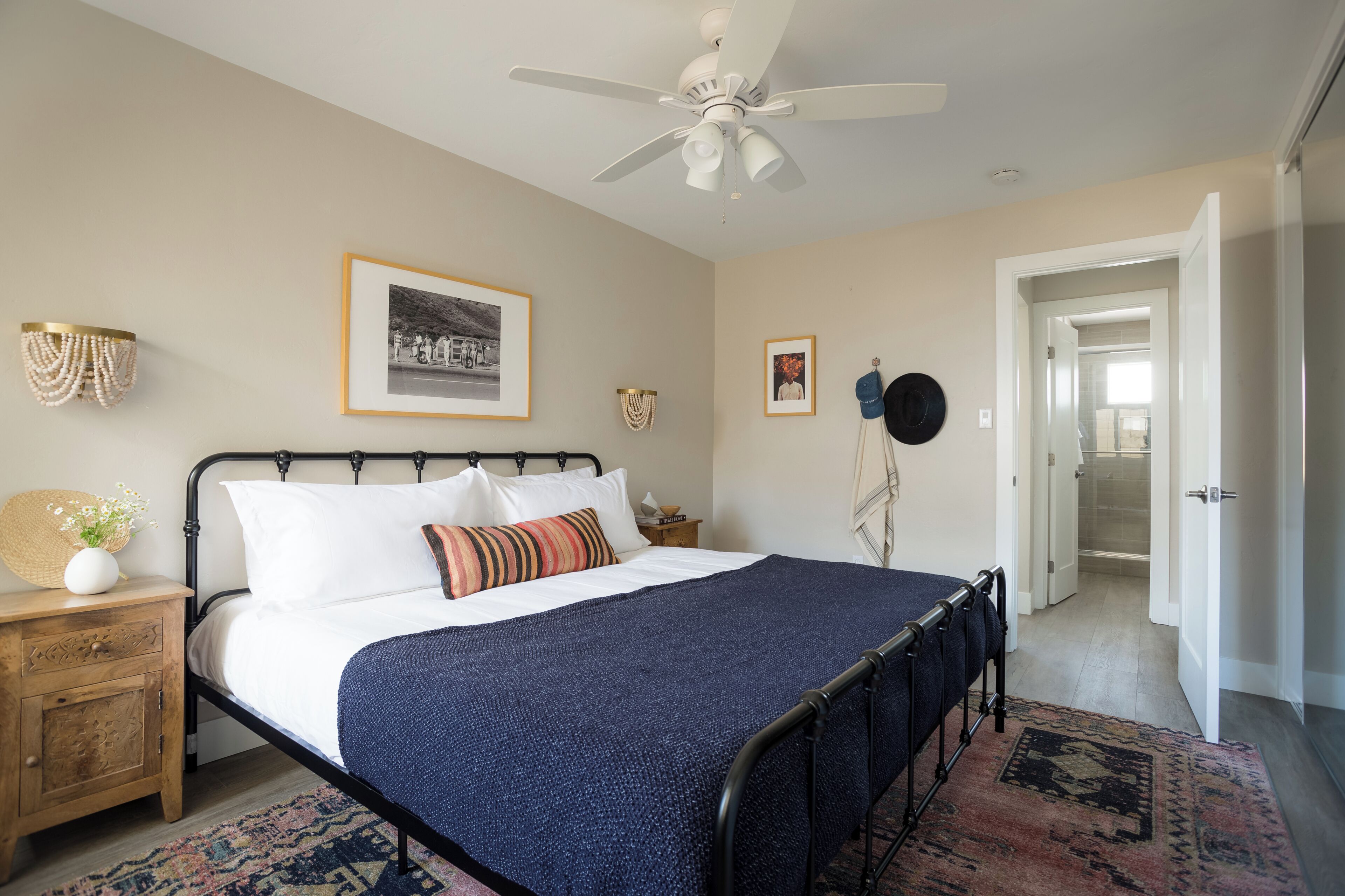 A bedroom with a bed, bedside tables, a ceiling fan, framed pictures, and a door leading to another room.