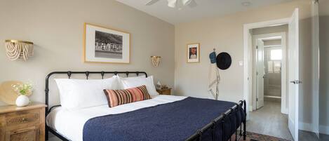 A bedroom with a bed, bedside tables, a ceiling fan, framed pictures, and a door leading to another room.