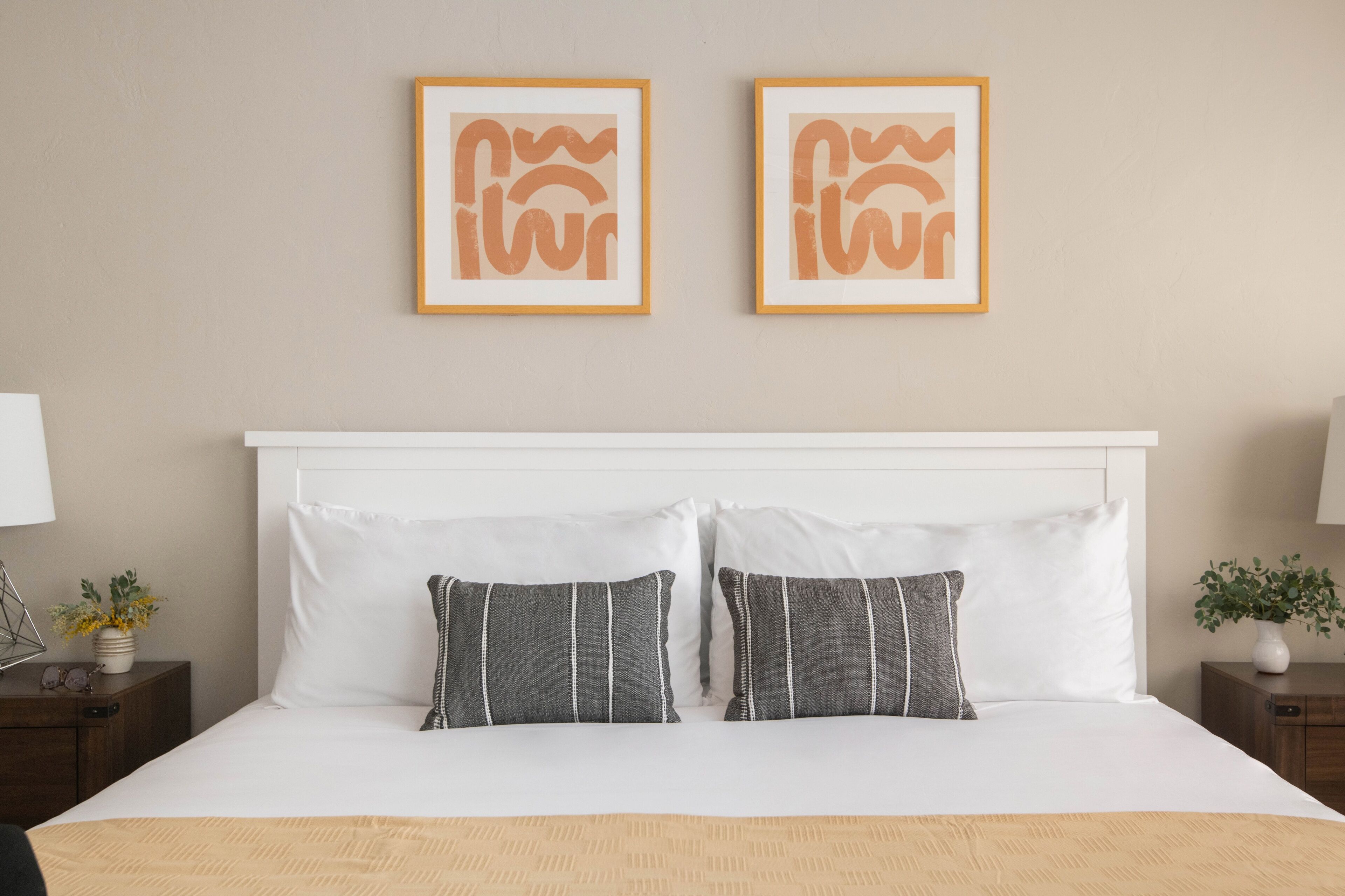 Una cama bien hecha con sábanas blancas y dos almohadas grises, un cabecero blanco y dos cuadros enmarcados en la pared.