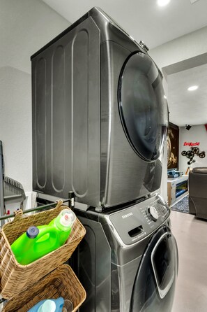 A modern laundry room with stacked washers and dryers, a wicker basket with cleaning supplies, and a wall-mounted projector. - Desert Hollywood UofA (Tucson)