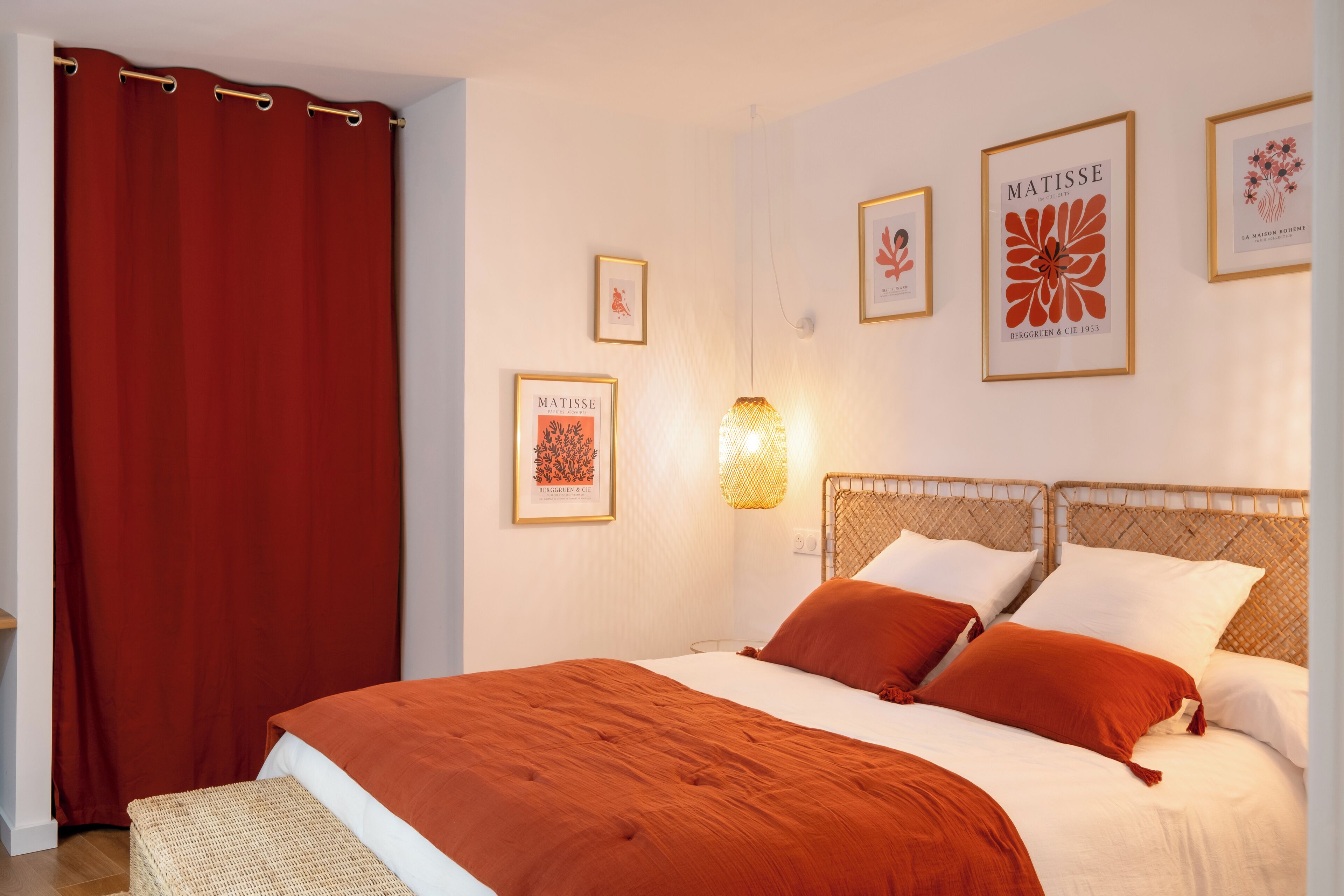 Une chambre à coucher bien aménagée, avec un grand lit, un rideau rouge et des œuvres d’art accrochées au mur, dont une œuvre de Matisse.
