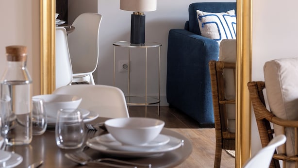 Un coin repas avec une table ronde dressée pour deux personnes, un miroir et un canapé bleu en arrière-plan.