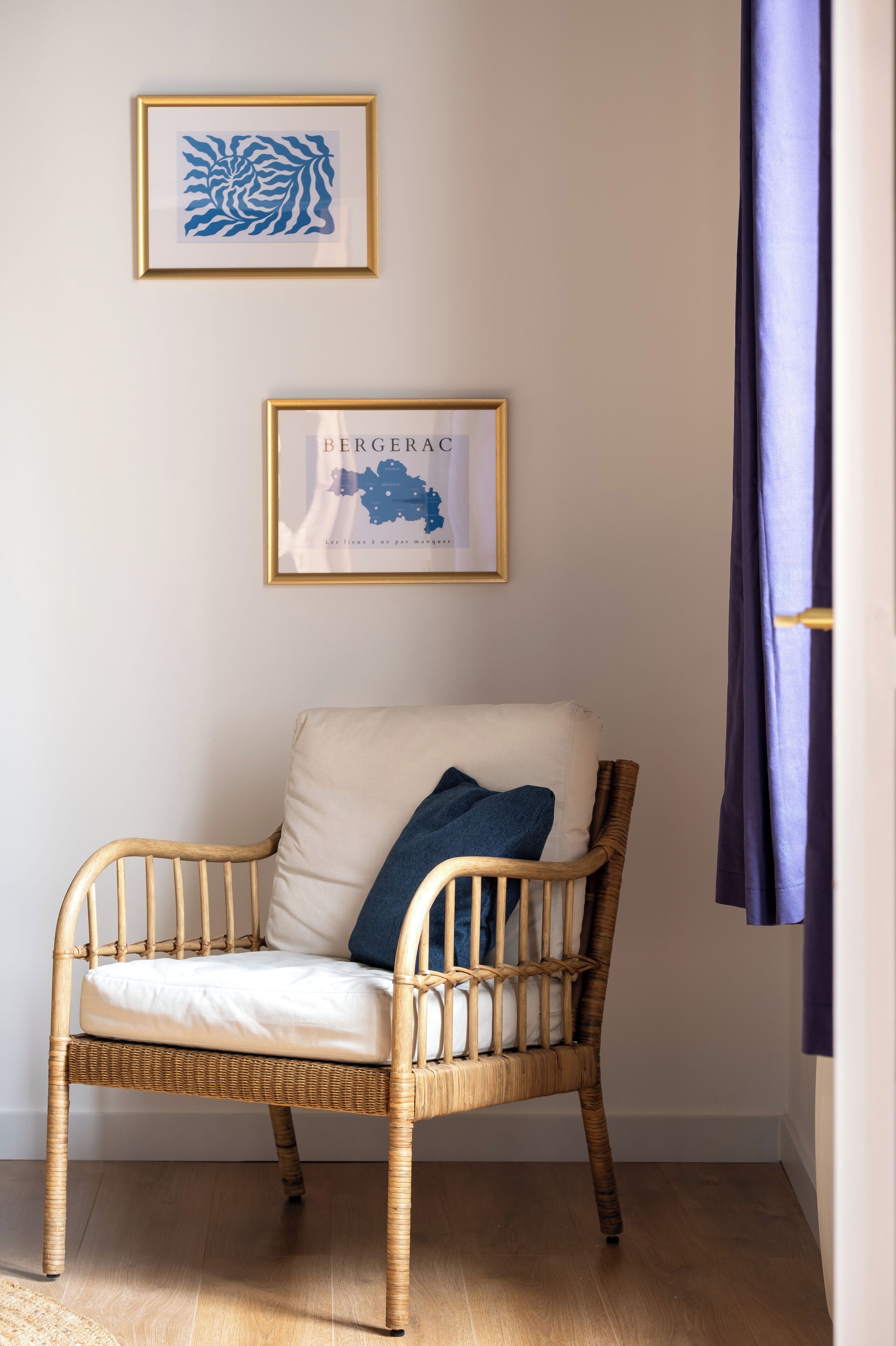 Une chaise en osier avec un coussin blanc et un oreiller bleu, placée contre un mur où sont accrochées deux œuvres d’art encadrées.
