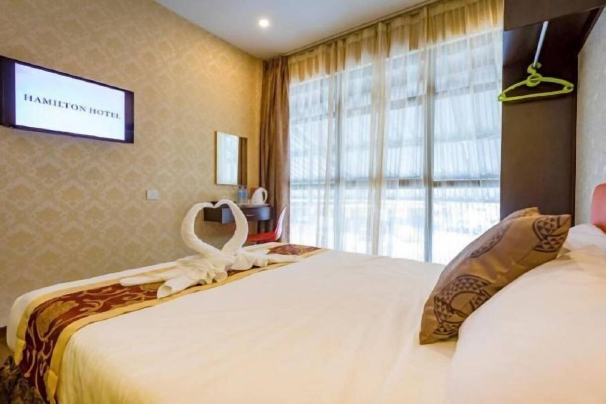 A hotel room with a bed, a heart-shaped towel arrangement, a mirror, and a window with curtains.