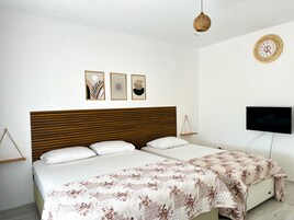 Un dormitorio con cabecera de madera, cama con ropa blanca, colcha estampada con motivos florales, televisor montado en la pared y tres obras de arte enmarcadas sobre la cama.