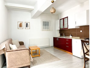 A compact living space with a sofa, a small wooden table, a kitchen area with red cabinets, and a radiator. - Şahan Apart (Yesilova)