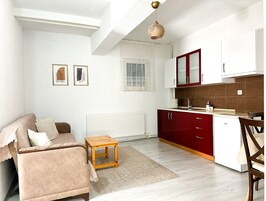 Un espacio habitable compacto con sofá, una pequeña mesa de madera, una zona de cocina con armarios rojos y un radiador.