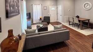 Ein modernes Wohnzimmer mit grauem Sofa, Holzböden, einer Essecke mit Holztisch und -stühlen sowie einer an der Wand befestigten Uhr.