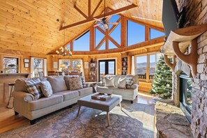 A spacious living room with a high ceiling, wooden beams, and large windows. There is a fireplace with a stone surround and a small Christmas tree.