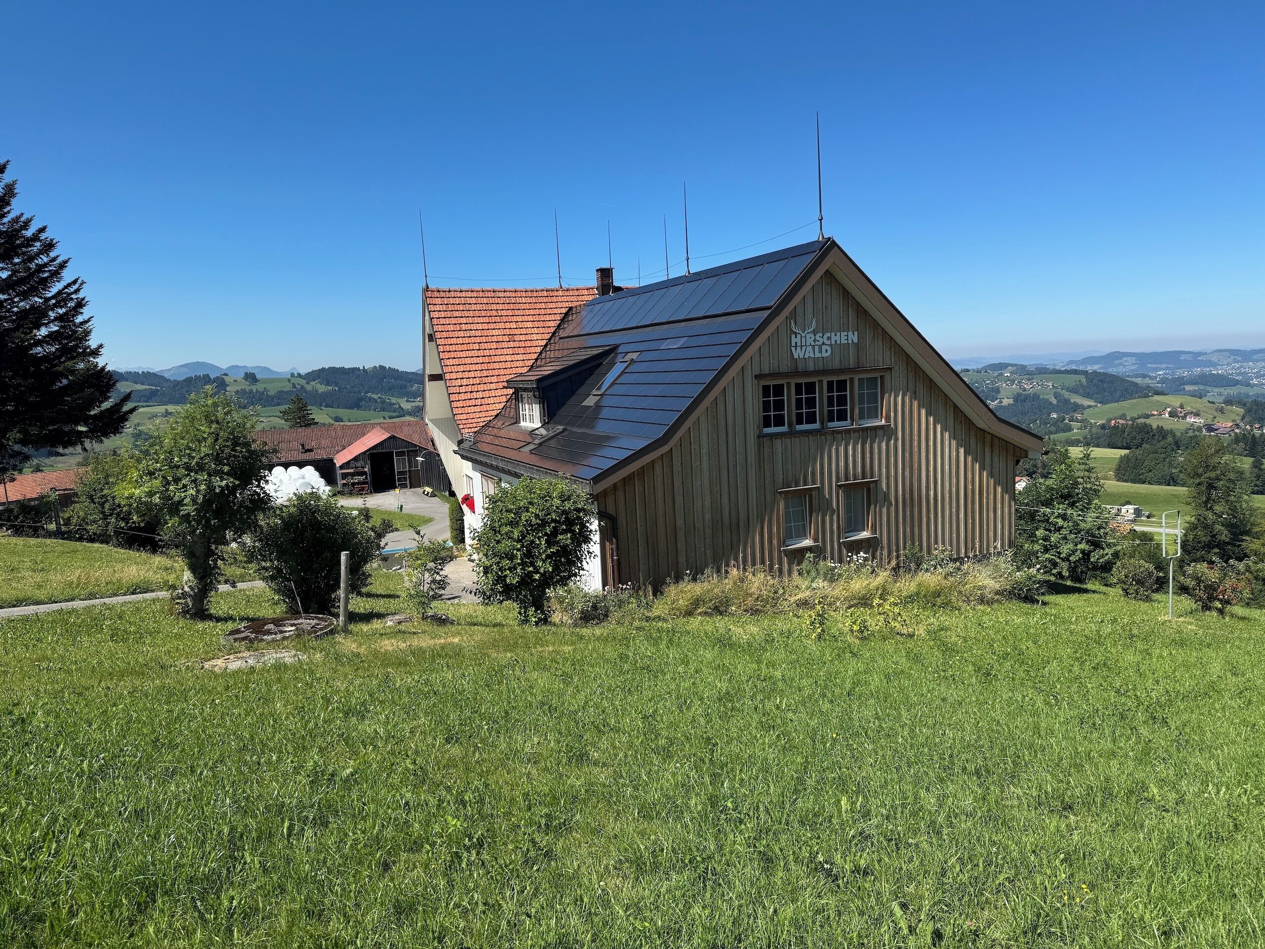 Ein hölzernes Gebäude mit geneigtem Dach, umgeben von einer grünen Wiese und Bäumen, mit Blick auf ferne Hügel und einen klaren Himmel.