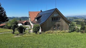 Ein hölzernes Gebäude mit geneigtem Dach, umgeben von einer grünen Wiese und Bäumen, mit Blick auf ferne Hügel und einen klaren Himmel.