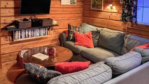 L’intérieur d’un chalet confortable, avec des murs en bois, un canapé gris, des coussins rouges et noirs, une petite table ronde sur laquelle reposent un livre et des lunettes, et une bibliothèque remplie de DVD.