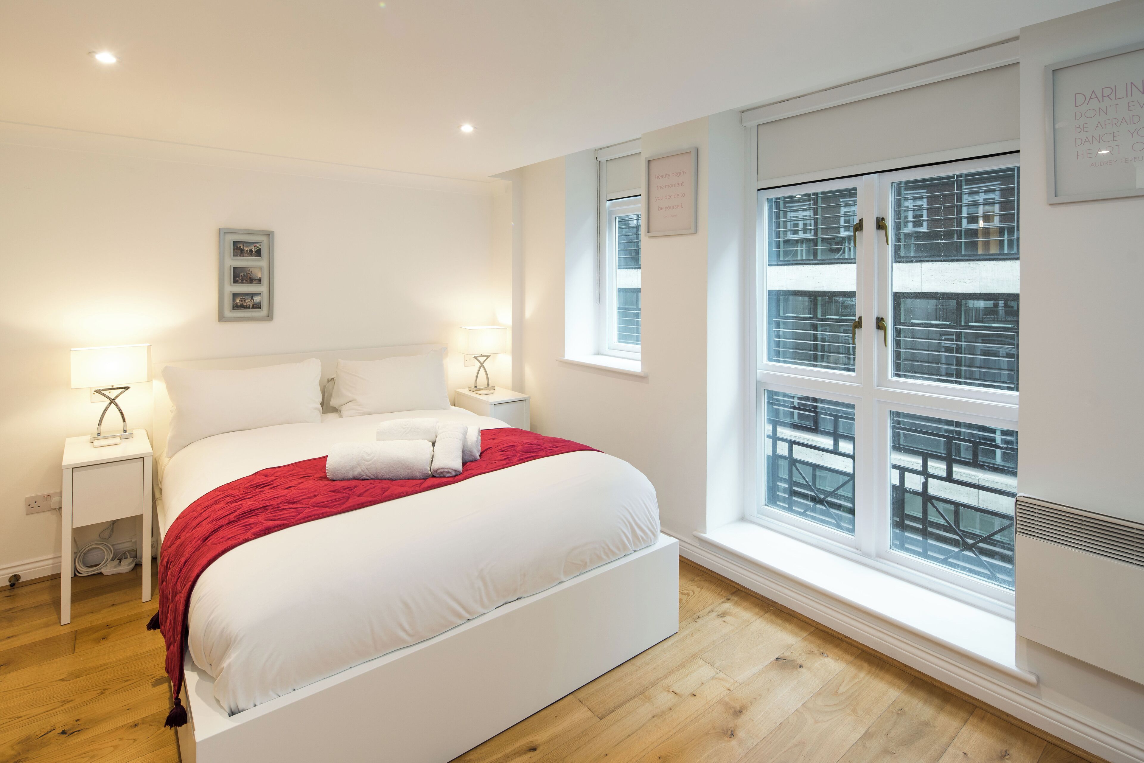Een moderne slaapkamer met een groot bed, witte beddengoed, een rode plaid, bedlampen en een raam met zonwering.