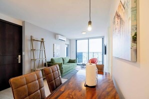 Una sala de estar moderna con una mesa de comedor, sillas de cuero, un sofá verde y un amplio ventanal con vistas a edificios.