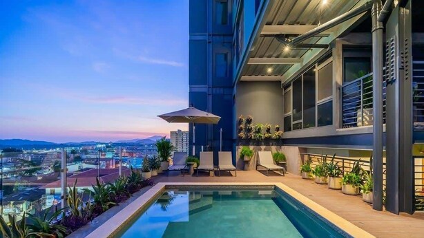 Piscina en la azotea con vistas a la ciudad, rodeada de sillones de descanso y plantas en macetas.