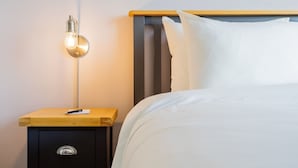Una cama bien tendida con sábanas blancas, una mesita de noche con lámpara y un pequeño aparador.