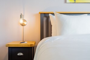A neatly made bed with white linens, a bedside table with a lamp, and a small drawer unit.