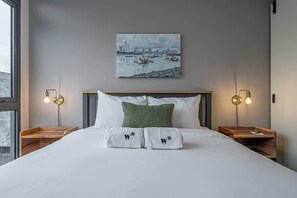 A hotel room with a large bed, two bedside tables with lamps, a cityscape painting on the wall, and a window.