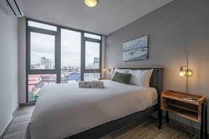 A hotel room with a large bed, a city view, a painting on the wall, and a bedside table with a lamp.