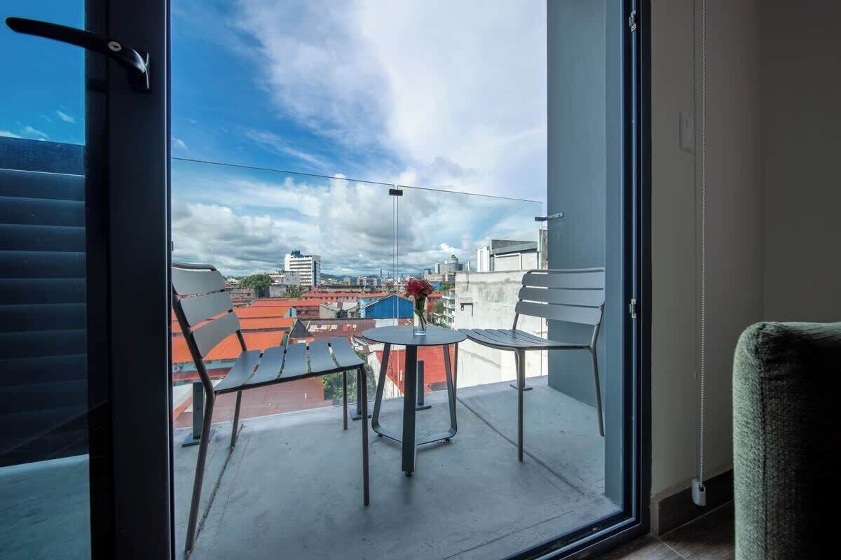 Un balcó amb taula i cadires, vista a la ciutat i una porta que dona a l'interior.