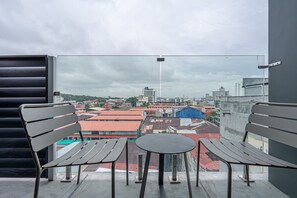 A balcony with two benches and a small table, overlooking a cityscape with buildings and a cloudy sky.