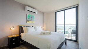 Une chambre d’hôtel avec un grand lit, une table de chevet, un appareil de climatisation et une vue sur l’extérieur par une grande fenêtre.